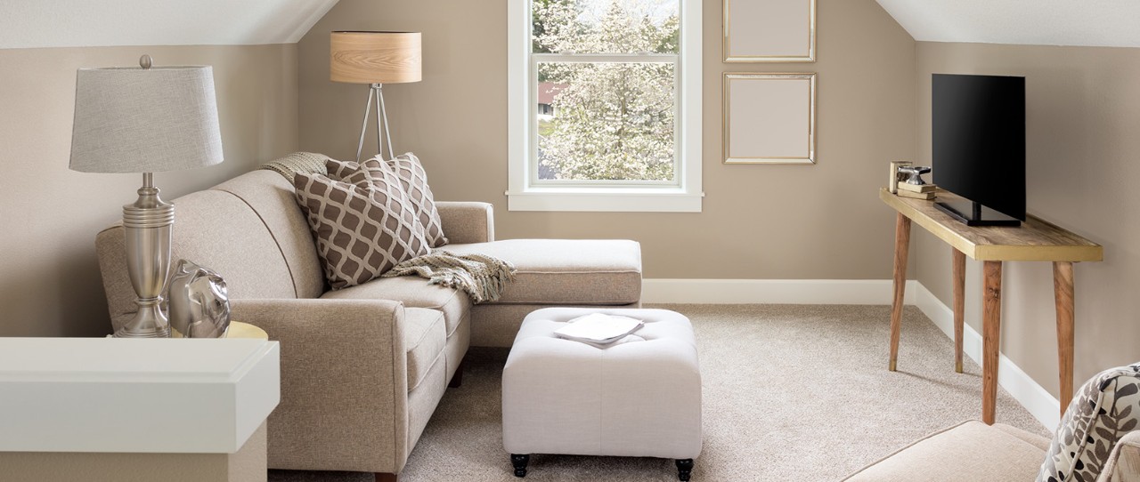 Salon bien éclairé par la lumière naturelle, avec un canapé beige avec chaise, un pouf blanc et une télévision.